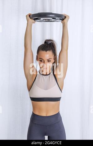 Jeune femme sportive forte tenant une plaque de poids lourde au-dessus de la tête tout en faisant des exercices de forme physique pendant l'entraînement à la maison Banque D'Images