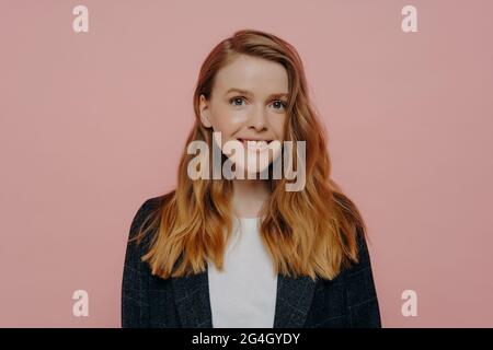 Jeune femme positive avec des cheveux de gingembre souriant à la caméra posant contre le mur rose Banque D'Images