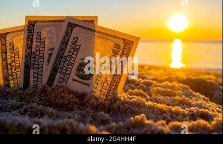 Trois billets de dollars sont enterrés dans le sable sur la plage de sable près de la mer à l'aube du coucher du soleil Banque D'Images