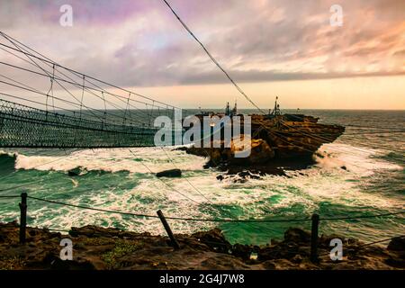 Défi sur le pont de pied de corde balançoire et le téléphérique à une île de roche avec de grandes vagues dangereuses à la plage de Pantai Timang, Java, Indonésie Banque D'Images