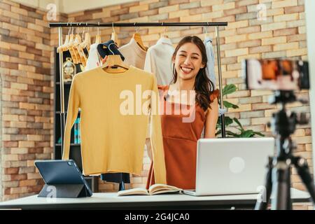 Jeune et belle femme asiatique blogueuse montrant des vêtements devant l'appareil photo de smartphone tout en enregistrant la vidéo vlog et la diffusion en direct dans sa boutique Banque D'Images