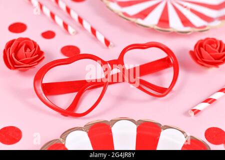 Verres en forme de cœur rouge, entourés de roses, confetti et assiettes en papier de fête sur fond rose Banque D'Images