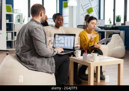 Homme d'affaires tenant un ordinateur portable avec des professionnels de la finance, tandis que divers employés parlent d'expliquer le plan de projet en examinant les documents administratifs assis sur le canapé. Collègues multiethniques analysant les rapports financiers Banque D'Images