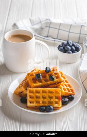 Gaufres belges avec baies et café sur une table en bois blanc. Boisson chaude du matin et myrtilles, concept petit déjeuner. Latte dans une tasse et pâtisserie sur un Banque D'Images