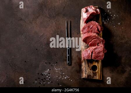 Filet de bœuf frais tranché de viande crue pour les steaks sur bois avec fourchette de viande en métal, sel, poivre sur fond brun foncé de texture. Cuisson des aliments b Banque D'Images