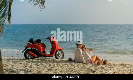 Trajet en scooter. Un couple charmant sur une moto rouge vêque de vêtements blancs sur une plage de sable. Les gens qui marchent près des palmiers tropicaux, de la mer. Location de motos. Banque D'Images