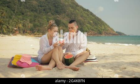 Le couple d'amour gai tenant et mangeant des tranches de pastèque sur le sable tropical plage mer. Amoureux romantiques deux personnes passent le week-end d'été. Banque D'Images