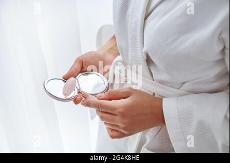 Image rognée d'une femme en peignoir blanc avec un rouleau de jade et un miroir dans ses mains. Gros plan des mains des femmes tenant un petit miroir de maquillage et un jade Banque D'Images