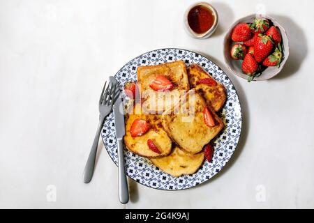 Un tas de toasts français avec des fraises fraîches sur une plaque en céramique, avec du sirop d'érable dans une carafe en céramique, une fourchette et un couteau sur fond de marbre blanc. Haut Banque D'Images