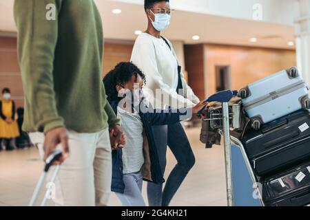 Famille à l'aéroport voyageant pendant la pandémie de Covid-19. Fille poussant le chariot à bagages tout en marchant avec les parents au terminal de l'aéroport. Banque D'Images