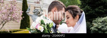 Vue latérale de la mariée avec le bouquet embrassant groom souriant près de la fontaine floue, bannière Banque D'Images
