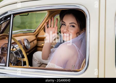 Une mariée souriante se fait la main à la caméra sur le siège du conducteur de la voiture vintage Banque D'Images