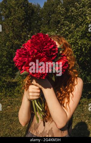 Jeune belle femme au gingembre bouclés tient des pivoines roses devant son visage. Banque D'Images