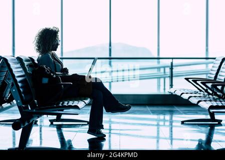 Une femme s'assoit seule à la porte de l'aéroport en attendant le vol pour voyager - voyage d'affaires ou de vacances - fenêtre de la vue lumineuse en arrière-plan - Banque D'Images