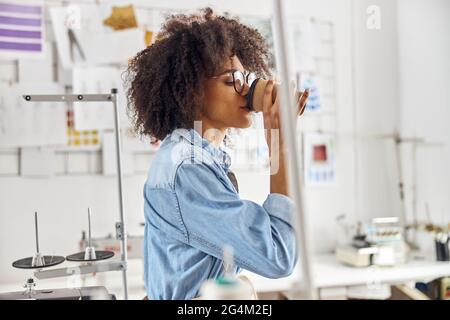 Une jeune femme afro-américaine en chemise de denim boit des boissons à l'aide d'équipements de couture Banque D'Images