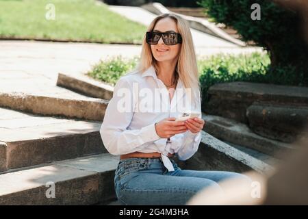 Femme élégante et gaie assise sur l'escalier dans le parc public, femme entrepreneur portant des lunettes de soleil et regardant loin avec Smile en attendant Banque D'Images