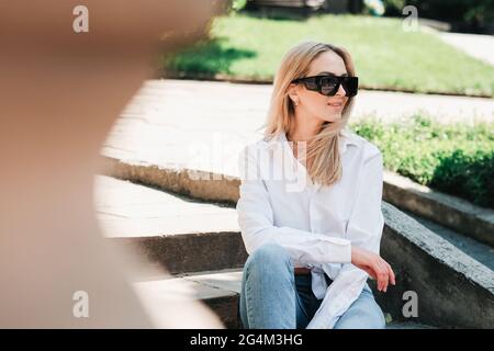 Femme élégante et gaie assise sur l'escalier dans le parc public, femme entrepreneur portant des lunettes de soleil et regardant loin avec Smile en attendant Banque D'Images