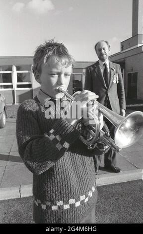 Années 1980, historique, à l'extérieur d'une école primaire, un jeune garçon jouant une trompette, un instrument de musique en laiton, avec son fier professeur de sexe masculin regardant sur, Angleterre, Royaume-Uni. Un son est fait par le trompettiste variant son mouvement de lèvre et souffle à travers un tube contrôlé par plusieurs valves. C'est en fait l'embout métallique qui produit le son. Banque D'Images