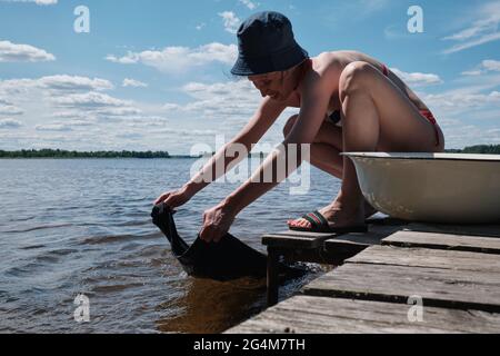 La jeune fille lave les vêtements à la main dans le lac. Laver à la main les vêtements dans la nature. Laver les vêtements dans le lac. Banque D'Images