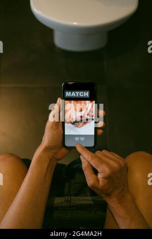 vue en grand angle d'un homme caucasien, assis dans les toilettes, datant en ligne dans son smartphone, avec une application simulée avec le mot match dans son écran Banque D'Images