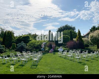 Cérémonie de mariage dans le magnifique jardin paysagé entre pelouse avec arc de mariage élégant décoré de fleurs fraîches et chaises blanches à côté. Banque D'Images