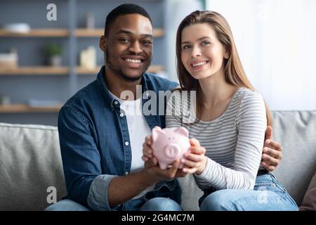 Joyeux jeune couple interracial tenant le Piggybank rose et souriant à la caméra Banque D'Images