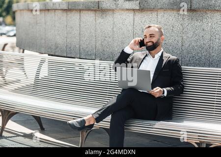 Homme d'affaires mûr avec ordinateur portable moderne assis sur un banc et parlant sur un smartphone, espace vide Banque D'Images