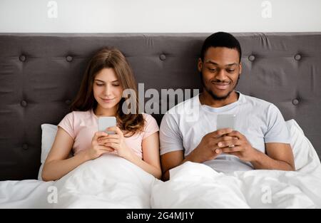 Un jeune couple multinational avec des téléphones portables au lit, se ignorant les uns les autres, coincé dans des jeux en ligne ou des réseaux sociaux Banque D'Images