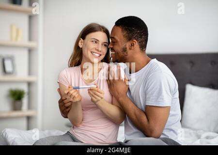 Couple multinational excité avec un test de grossesse positif se portant sur le lit, célébrant la réussite de la grossesse à la maison Banque D'Images