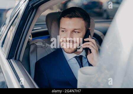 Image d'un jeune juriste caucasien avec succès dans un costume bleu foncé formel parlant sur un téléphone portable en voiture Banque D'Images