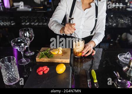 Courte femme barkeeper non reconnaissable avec une tenue élégante ajoutant des glaçons dans le verre et remuant avec une longue cuillère tout en préparant un cocktail debout Banque D'Images