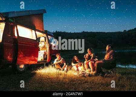 Groupe d'amis joyeux ayant du camping amusant près du lac et de la camionnette pendant la nuit étoilée Banque D'Images