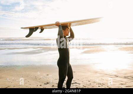 Vue latérale d'un surfeur vêtu d'une combinaison de plongée qui marche sur la plage avec la planche de surf au-dessus de sa tête le matin et le lever du soleil en arrière-plan Banque D'Images