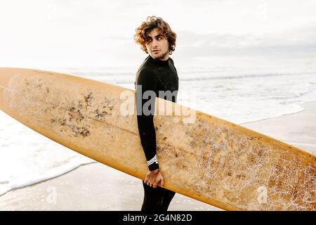 Vue latérale d'un surfeur vêtu d'une combinaison de plongée en regardant loin avec la planche de surf sur la plage pendant le lever du soleil en arrière-plan Banque D'Images