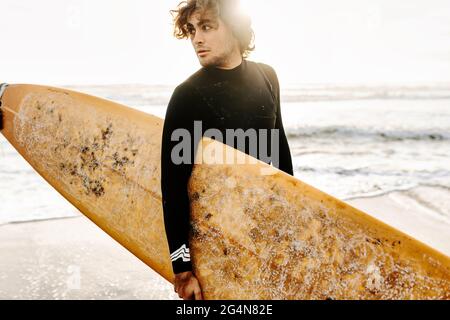 Vue latérale d'un surfeur vêtu d'une combinaison de plongée en regardant loin avec la planche de surf sur la plage pendant le lever du soleil en arrière-plan Banque D'Images