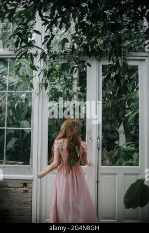 Vue arrière d'une femelle méconnaissable avec de longs cheveux de gingembre atteignant la main à la porte de la serre avec des plantes Banque D'Images