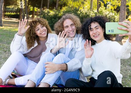 Groupe de jeunes femmes et hommes multiraciaux avec des cheveux bouclés assis sur l'herbe verte dans le parc et en agitant les mains tout en prenant le selfie avec le téléphone mobile Banque D'Images