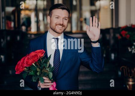 Jeune homme beau avec une barbe nette dans un smoking élégant avec des roses rouges dans sa main Banque D'Images