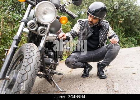 Corps complet de cycliste adulte de race masculine dans une tenue élégante et un casque assis sur des haunches et de vérifier les détails de moto cassée sur la route près de Lush Banque D'Images