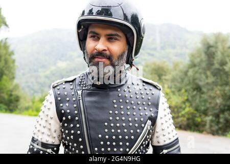 Un homme hispanique d'âge moyen positif, portant une veste en cuir tendance avec rivets et casque noir souriant et regardant loin en étant debout sur la route Banque D'Images