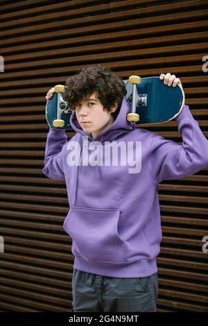 Adolescent sans émotion dans des vêtements décontractés regardant l'appareil photo tout en se tenant debout avec le skateboard derrière la tête sur la rue avec un arrière-plan flou Banque D'Images