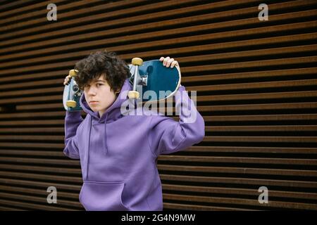 Adolescent sans émotion dans des vêtements décontractés regardant l'appareil photo tout en se tenant debout avec le skateboard derrière la tête sur la rue avec un arrière-plan flou Banque D'Images