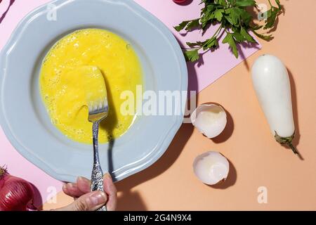 Vue de dessus de la récolte femelle méconnaissable battant des œufs non cuits contre des brins de persil frais et de l'aubergine blanche sur fond de deux couleurs Banque D'Images