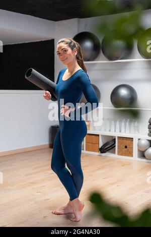 Corps complet de la tenue pieds nus jeune femme en vêtements de sport avec tapis de yoga roulé dans la main souriant et regardant l'appareil photo tout en préparant pour le cours de fitness s Banque D'Images