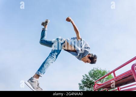 Jeune homme intrépide et bas angle sautant au-dessus de la rampe métallique en ville tout en effectuant un spectacle de cascades par temps ensoleillé Banque D'Images