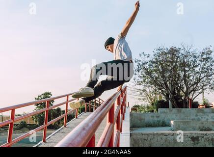 Jeune homme intrépide et bas angle sautant au-dessus de la rampe métallique en ville tout en effectuant un spectacle de cascades par temps ensoleillé Banque D'Images