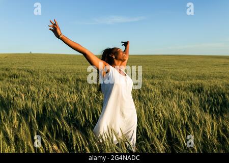 Jeune femme noire souriante en robe d'été blanche se promenant sur le champ de blé vert tandis que les yeux fermés dans la journée sous le ciel bleu Banque D'Images