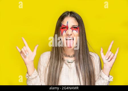 Jeune femme élégante avec des cornes de maquillage créatives et montrant la langue sur fond jaune Banque D'Images