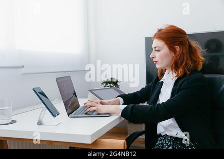 Vue latérale d'une jeune entrepreneur travaillant sur un netbook au bureau avec une tablette et un smartphone à la maison Banque D'Images