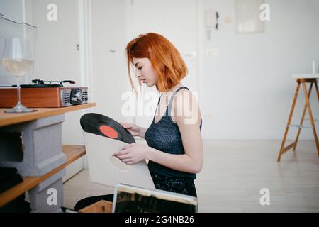 Jeune femme sélectionnant le disque en vinyle dans un conteneur en bois tout en étant assise contre la plaque tournante d'époque dans la chambre Banque D'Images